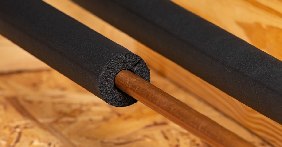 A close-up of two pipes fitted with black foam insulation sleeves resting on a wooden subfloor surface.