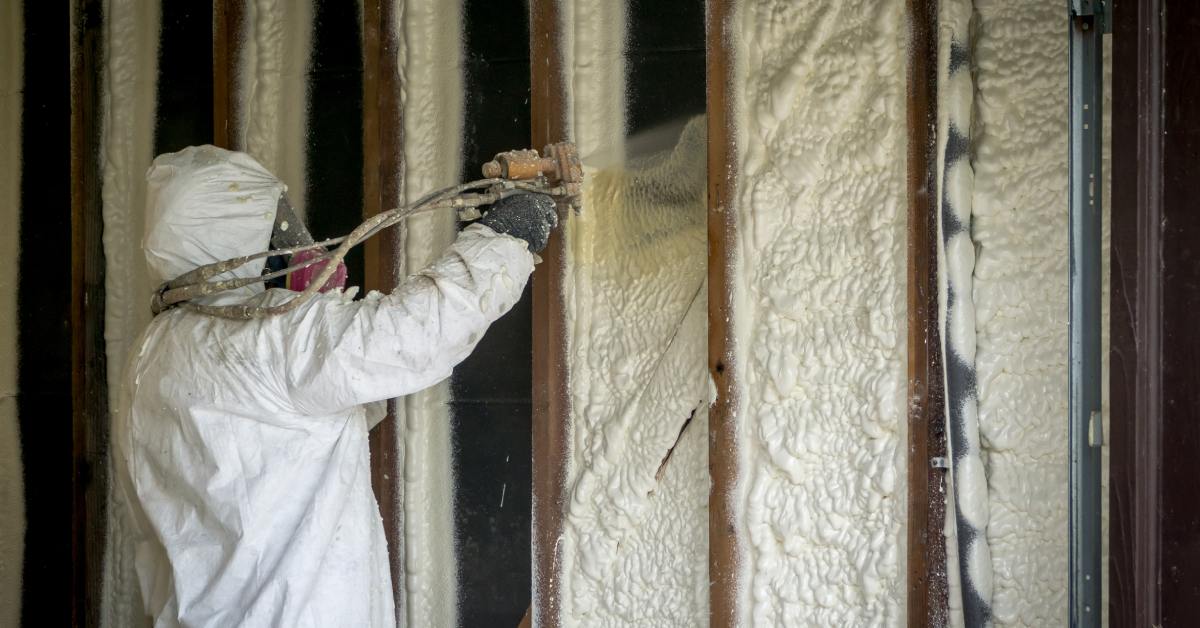 A person with professional gear sprays closed-cell spray foam insulation between the wooden beams of a wall.