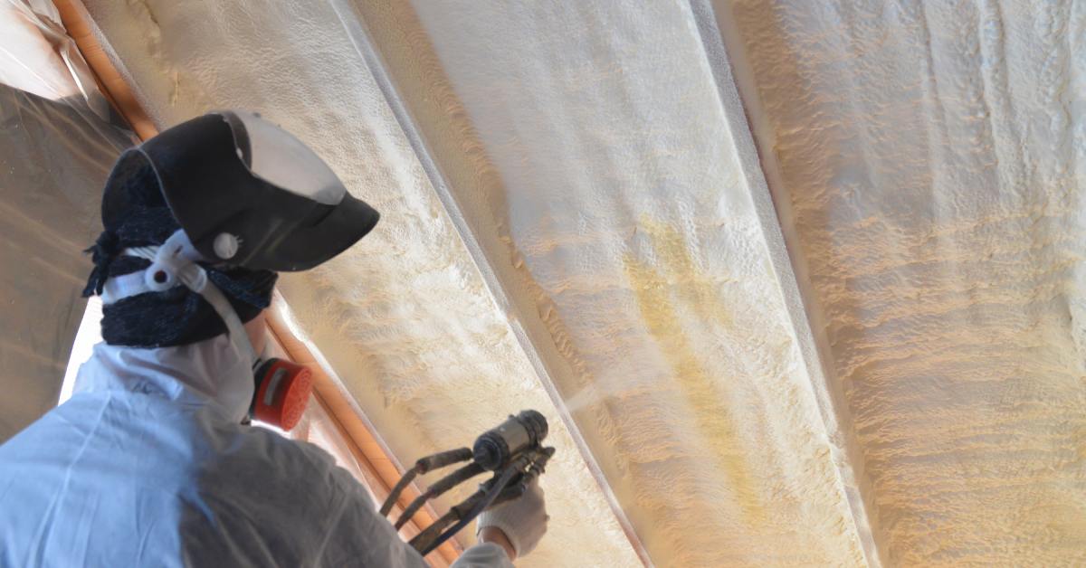 A person wearing a safety mask and suit uses a spray gun to apply foam insulation on the crevices of a roof.