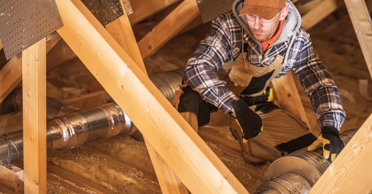 A worker installing HVAC ductwork in a cold attic space during winter, surrounded by insulation and wooden beams.