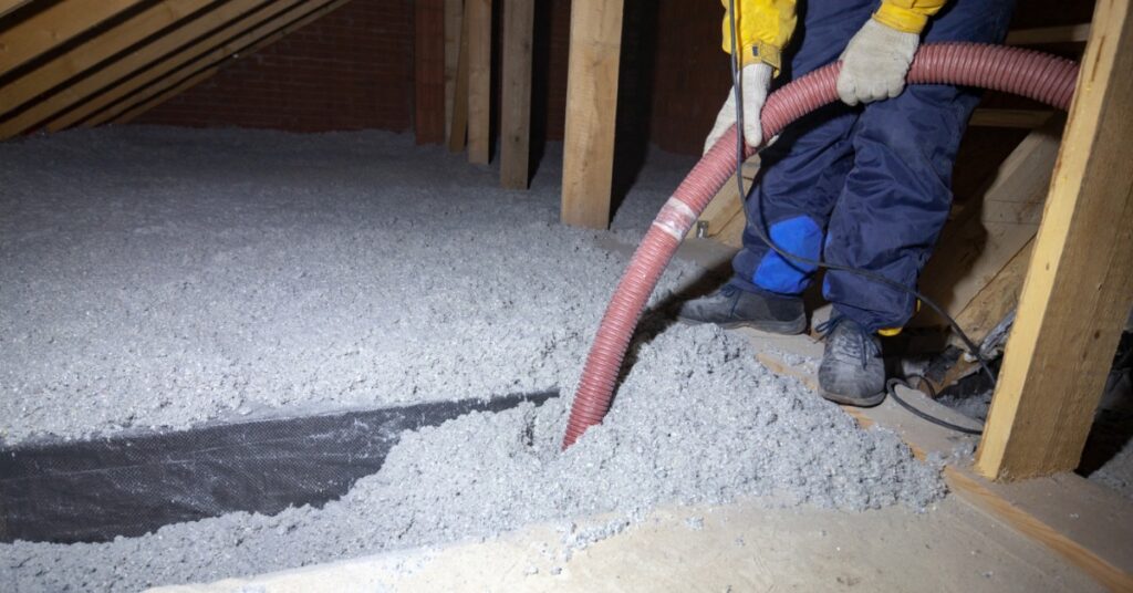 a person wearing work clothes holds a red hose while loose insulation covers the floor of a timber attic.
