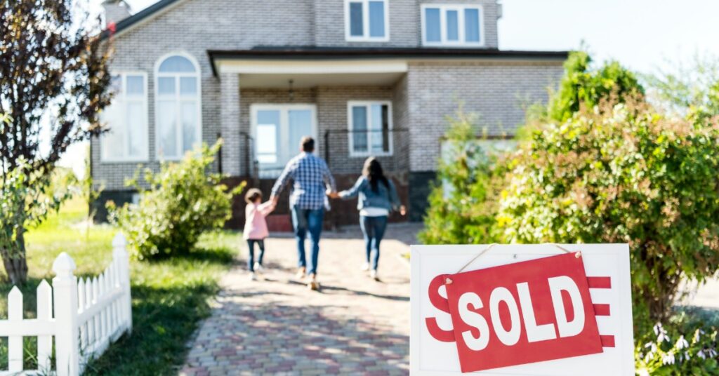 a family walks toward a brick house along a paved path while a red sold sign stands beside green shrubs.