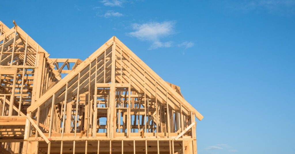 A wooden house structure with open trusses and gabled rooflines stands under a bright blue sky with a few clouds.