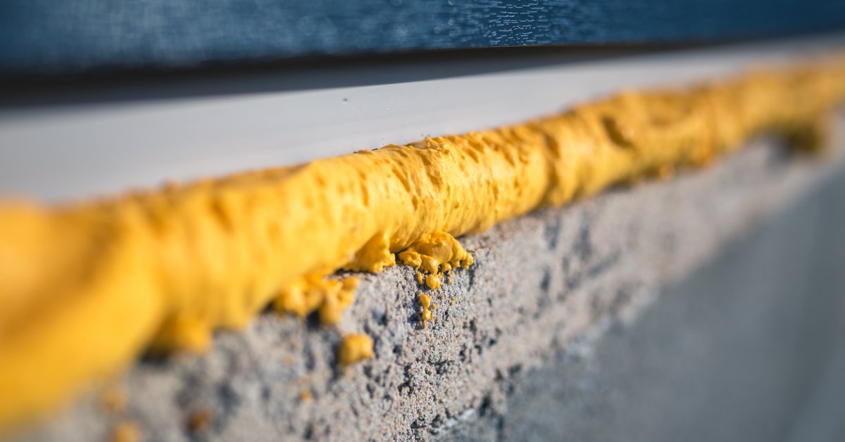 A close-up of yellow expanding foam filling a gap between a rough concrete edge and a blue window surface.