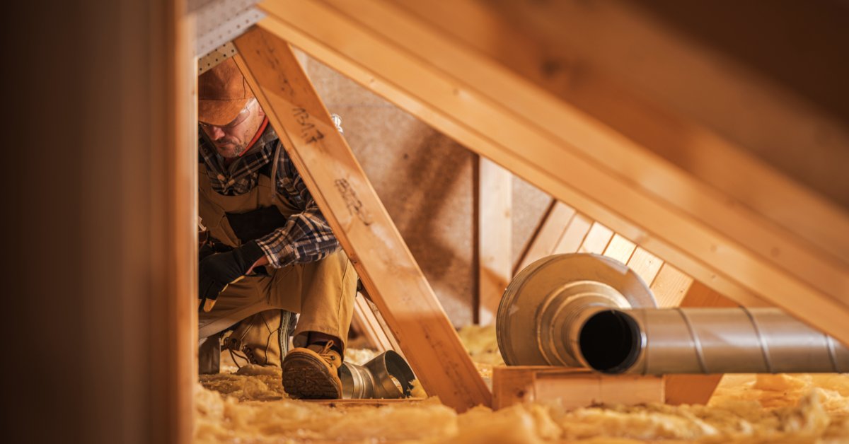 A person working in an attic near wooden beams, insulation, and metal ventilation ducts under warm interior light.