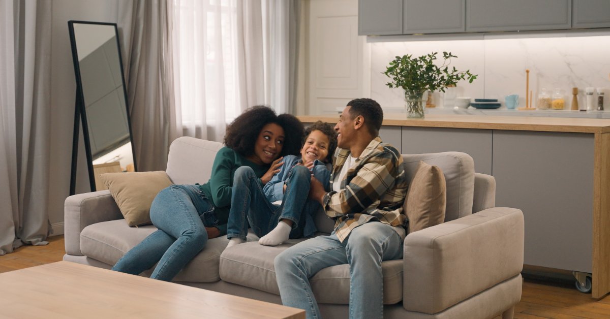 Two adults and a child sitting together on a grey couch in a bright living room, smiling at one another.