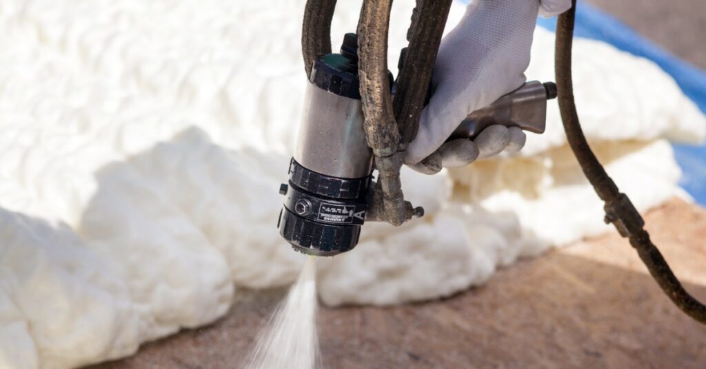 A person in protective gear sprays foam insulation between wooden wall studs with a hose and spray gun.