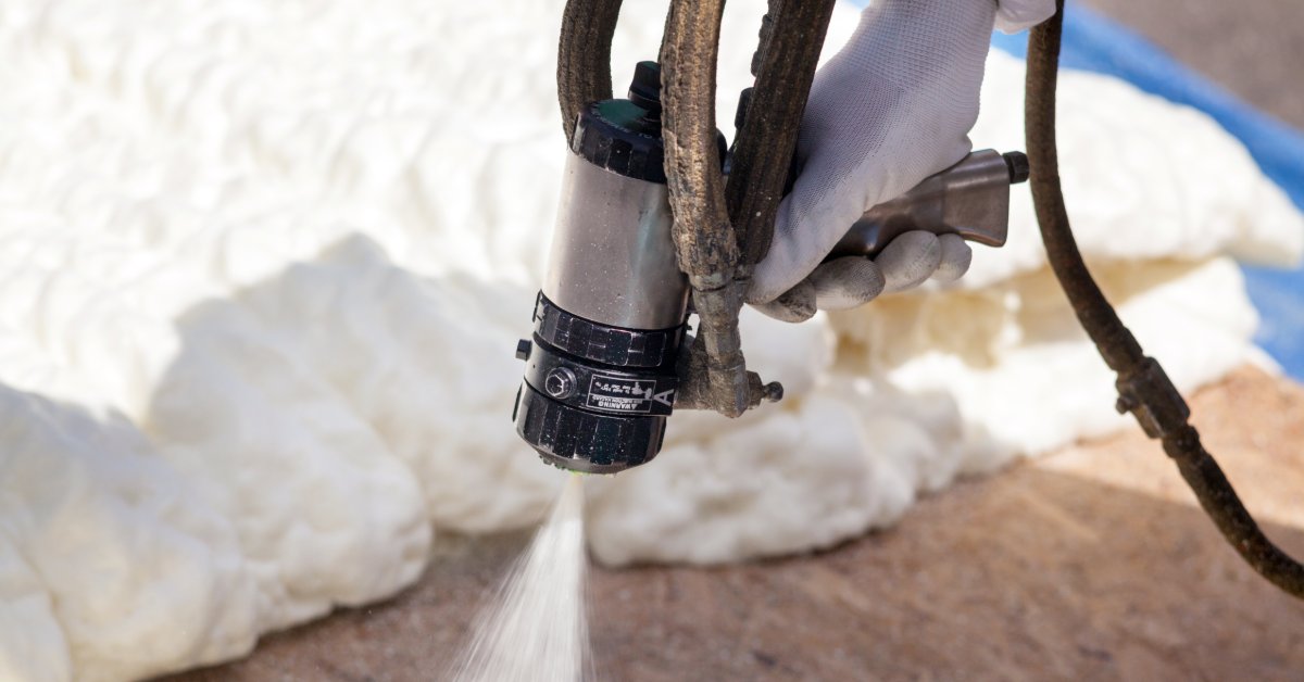 A gloved hand holding a spray foam gun over white insulation on a wooden surface, with attached hoses above.