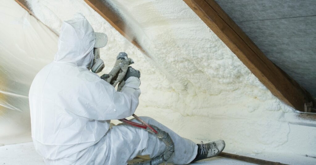A person in a white protective suit spraying foam insulation between wooden roof beams inside an attic.