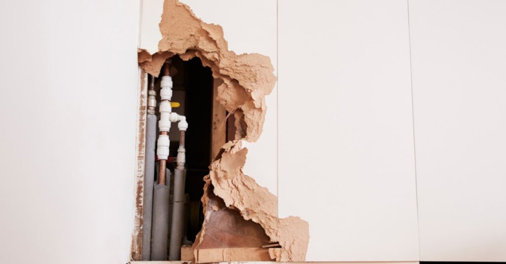 A damaged interior wall exposing pipes, torn drywall, and wooden framing above a tiled floor inside a room.