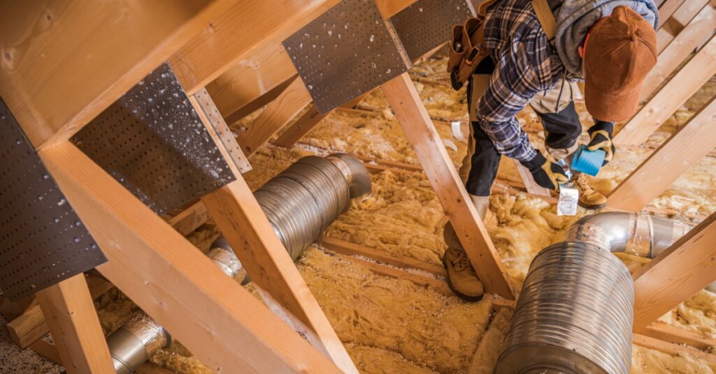 A worker applying tape near silver ventilation ducts and blown-in cellulose between wooden attic beams.