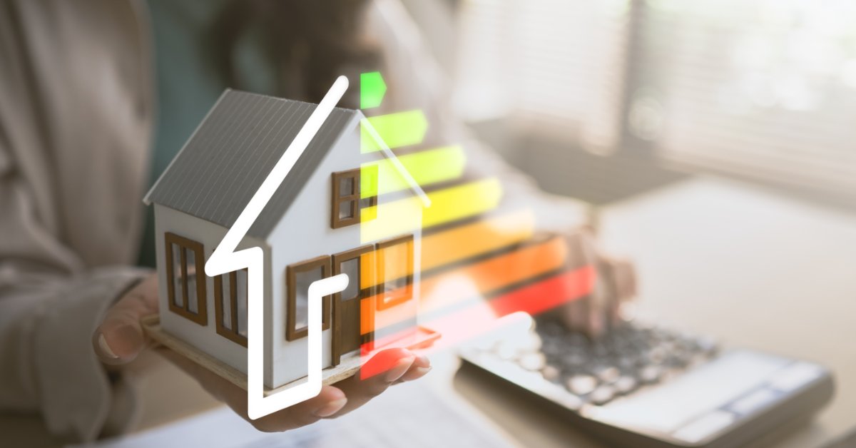 A person holds a small house model next to a colorful energy-rating graphic over a laptop and paperwork.