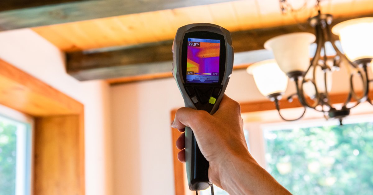 A person points a thermal scanner toward wooden ceiling beams and light fixtures inside a room with windows.