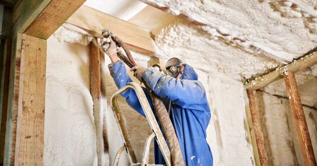 A person in blue protective gear sprays foam insulation along a wall-ceiling junction inside a framed home.