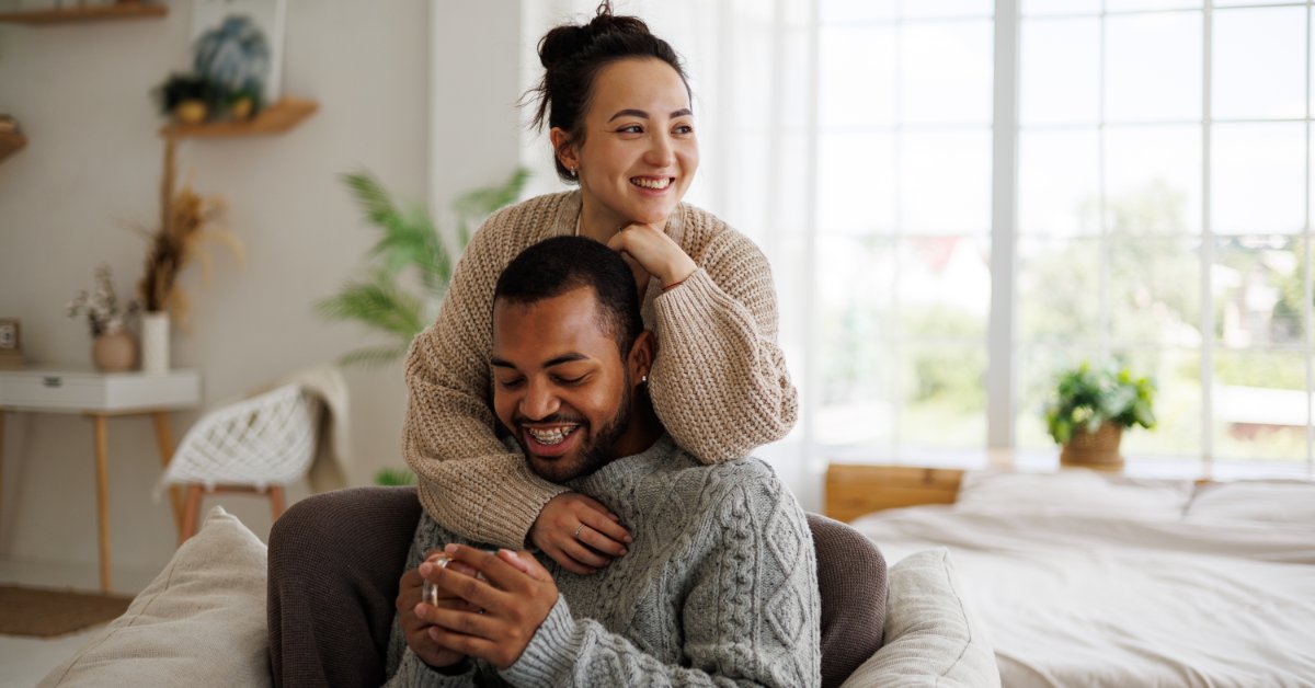 Two people wearing sweaters sit together on a couch indoors in soft lighting. One person holds a mug and smiles.