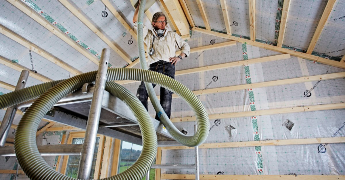A worker stands on a platform, installing blown cellulose insulation inside an attic using a flexible hose.