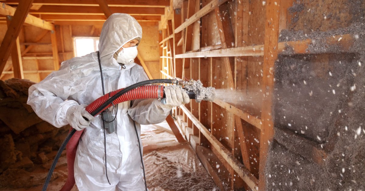 A close-up of a worker in a protective suit spraying cellulose insulation into an exposed wall cavity.