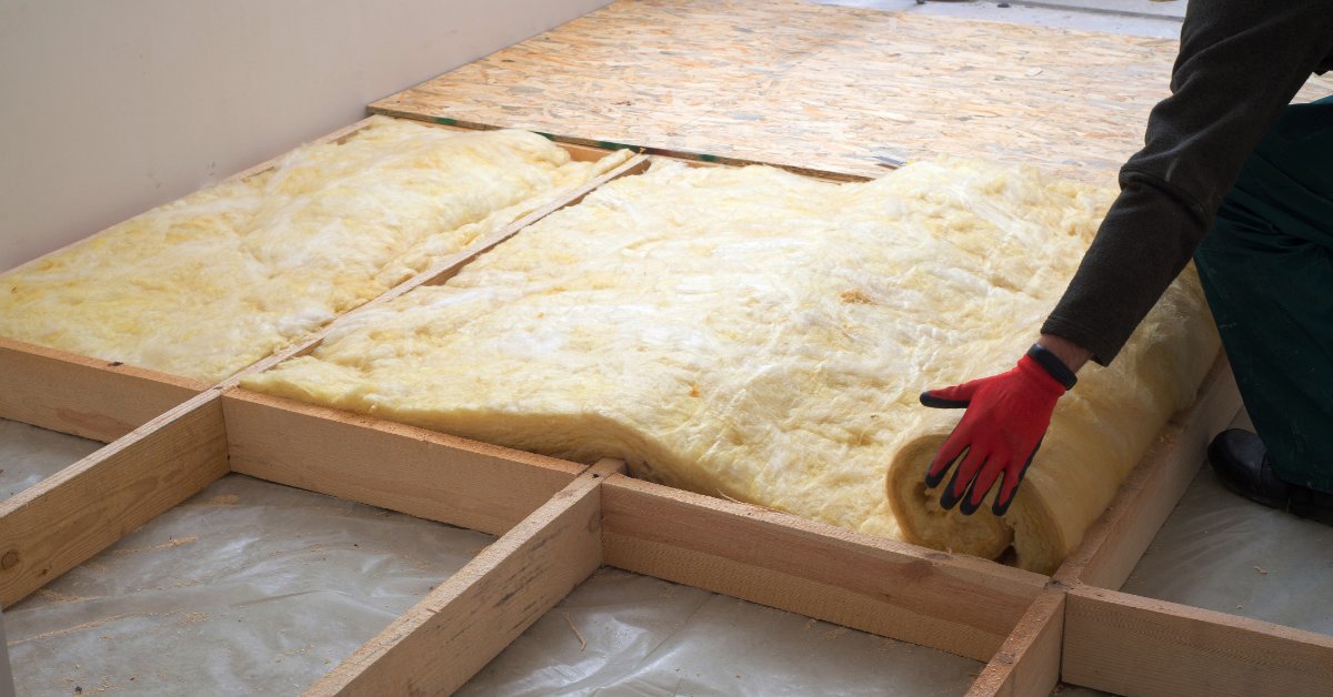 A worker wearing red gloves rolling yellow mineral wool insulation between exposed wooden floor joists in a room.