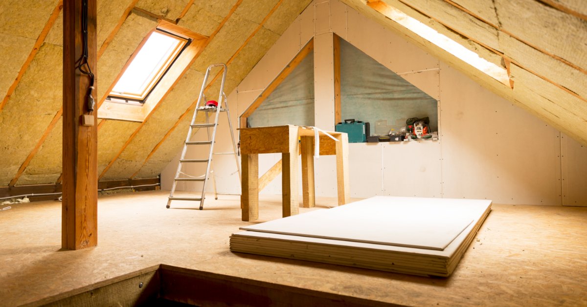 An unfinished attic with sloped insulated ceilings, skylights, a ladder, and stacked white panels on the wooden floor.
